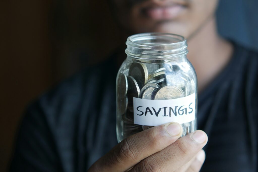 Ein Mann hält ein Sparglas mit der Aufschrift Savings voller Münzen in der Hand, um Geld für die Reiseapotheke Checkliste und wichtige Medikamente anzusparen.