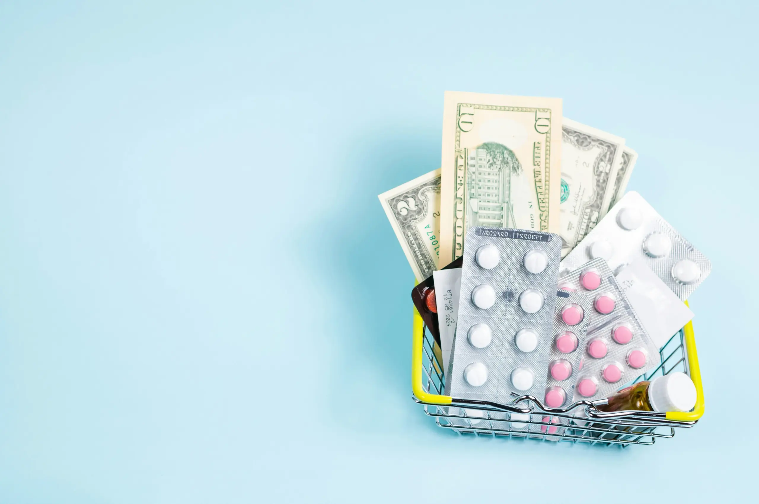 Ein kleiner Einkaufskorb gefüllt mit Medikamenten-Blistern, Pillen und US-Dollar-Geldscheinen auf blauem Grund als Symbol für die Kosten einer Basis Reiseapotheke.