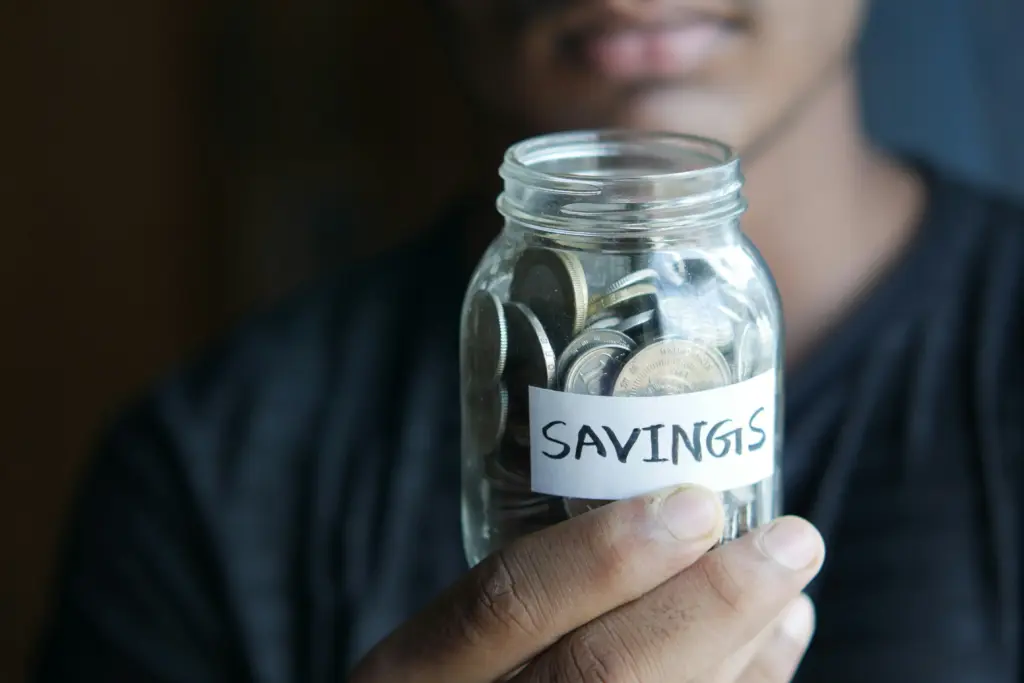 Ein Mann hält ein Sparglas mit der Aufschrift Savings voller Münzen in der Hand, um Geld für die Reiseapotheke Checkliste und wichtige Medikamente anzusparen.