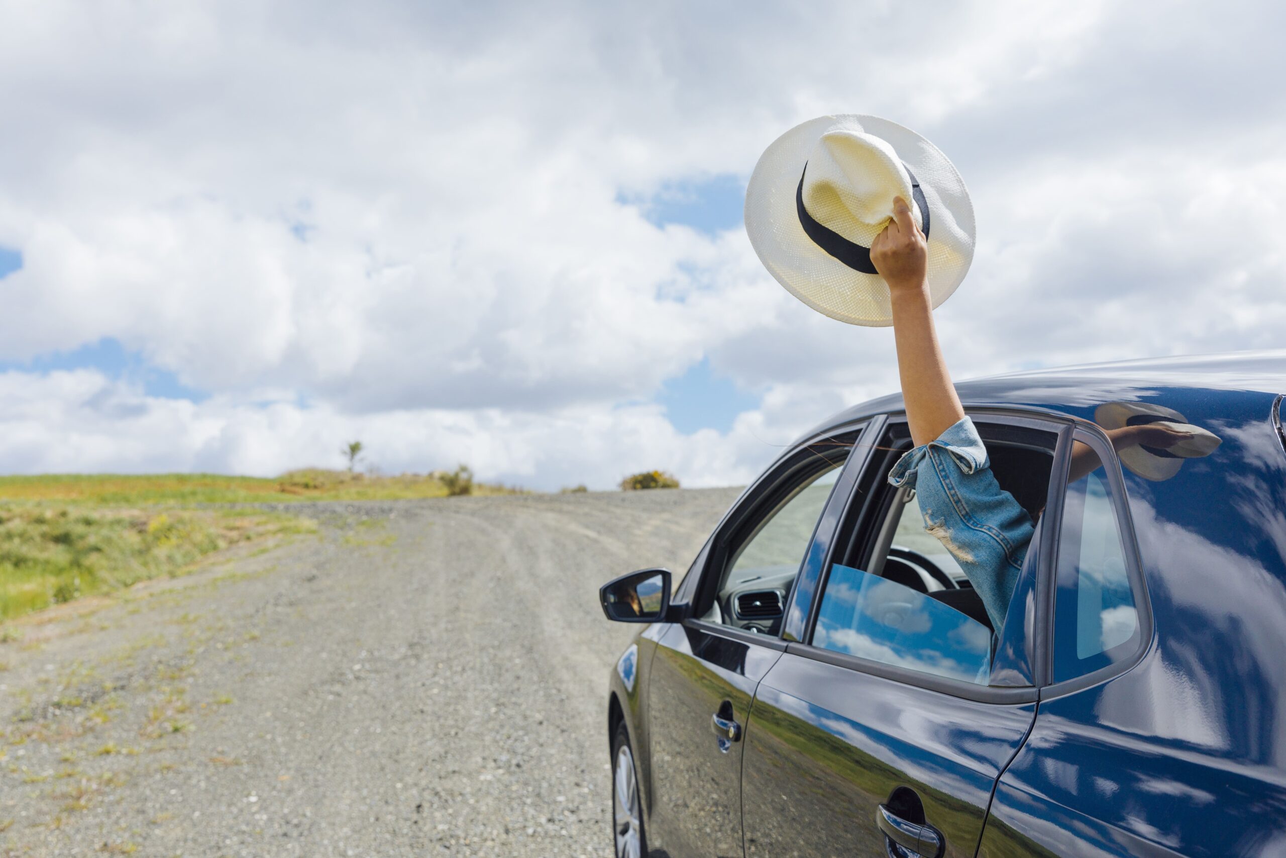 Eine Nahaufnahme eines blauen Autos auf einem Schotterweg, von dessen Beifahrerseite ein Arm mit einem Hut in die Luft geschwenkt wird. Der Arm ist in eine Jeansjacke gekleidet, und der Hut ist ein weißer Strohhut mit einem schwarzen Band. Der Schotterweg führt in eine grüne Landschaft mit sanften Hügeln und einer großen, flachen Grasfläche. Der Himmel ist hellblau mit lockeren weißen Wolken. Das Foto ist aus der Perspektive eines stehenden Beobachters aufgenommen, der dem Auto auf dem Schotterweg folgt.