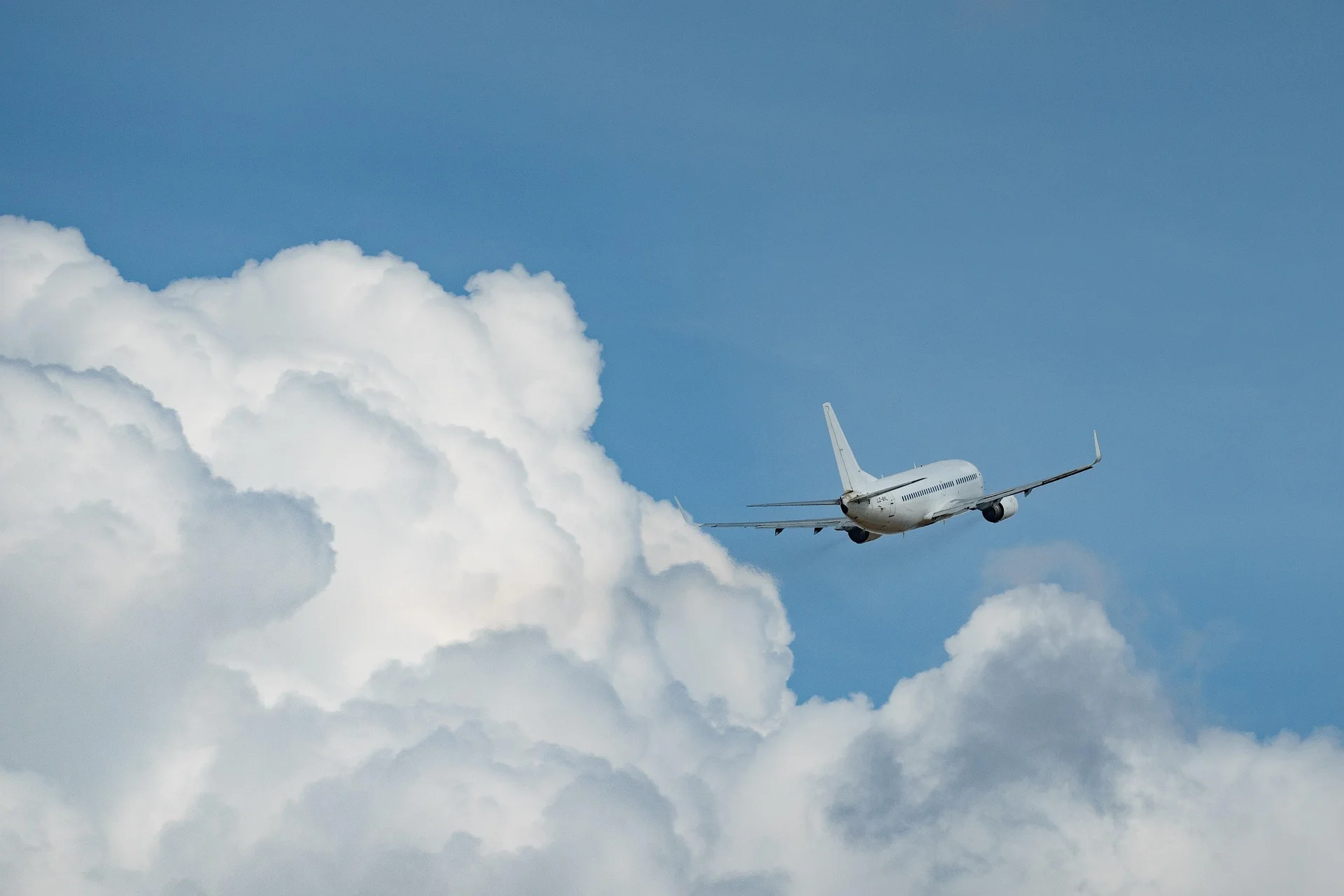 Die Rückansicht eines weißen Verkehrsflugzeugs beim Steigflug in einen klaren, blauen Himmel mit einer großen, weißen Kumuluswolke auf der linken Seite. Das Flugzeug hebt sich vom blauen Hintergrund ab und fliegt nach rechts oben.