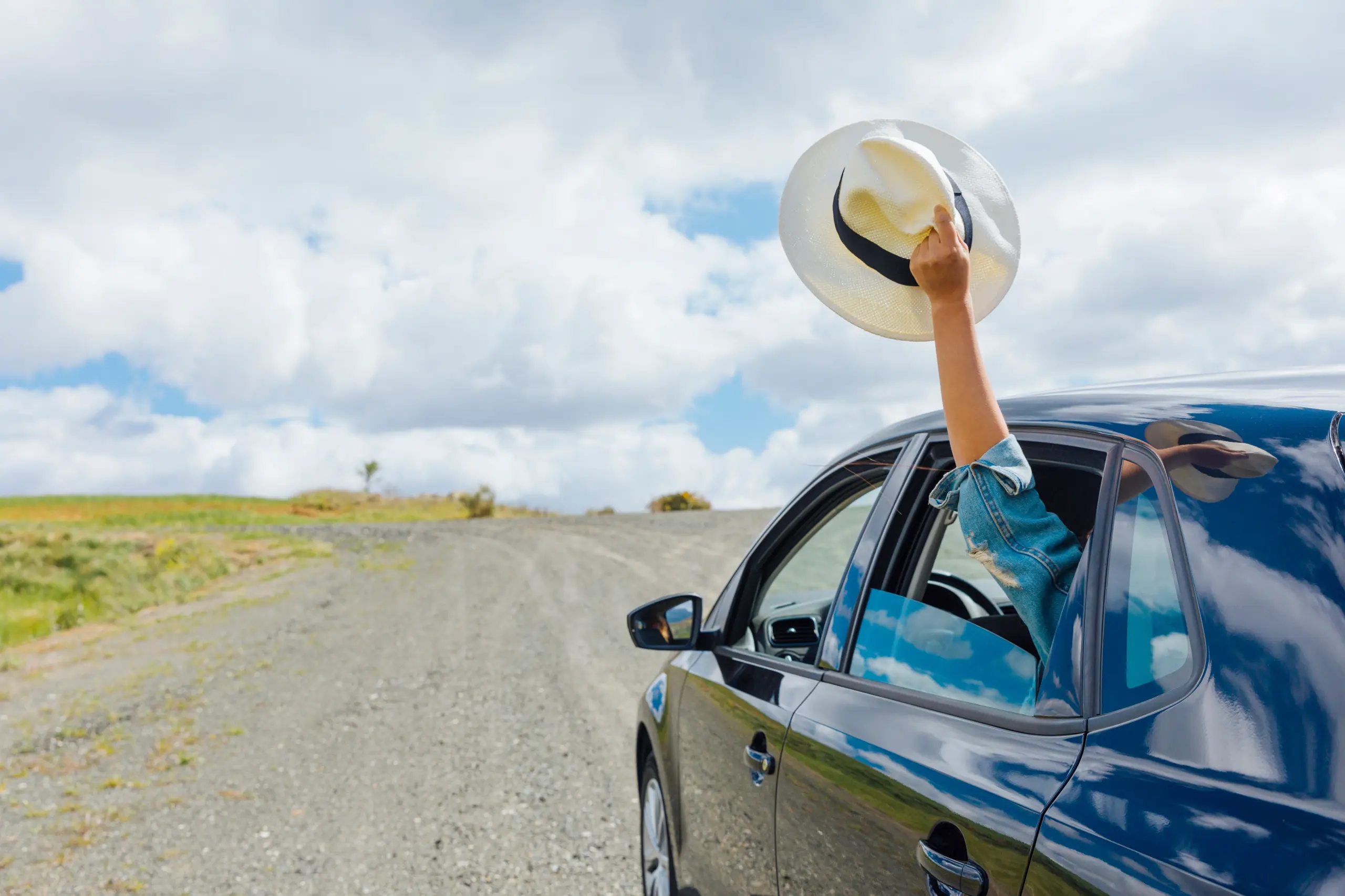 Eine Nahaufnahme eines blauen Autos auf einem Schotterweg, von dessen Beifahrerseite ein Arm mit einem Hut in die Luft geschwenkt wird. Der Arm ist in eine Jeansjacke gekleidet, und der Hut ist ein weißer Strohhut mit einem schwarzen Band. Der Schotterweg führt in eine grüne Landschaft mit sanften Hügeln und einer großen, flachen Grasfläche. Der Himmel ist hellblau mit lockeren weißen Wolken. Das Foto ist aus der Perspektive eines stehenden Beobachters aufgenommen, der dem Auto auf dem Schotterweg folgt.